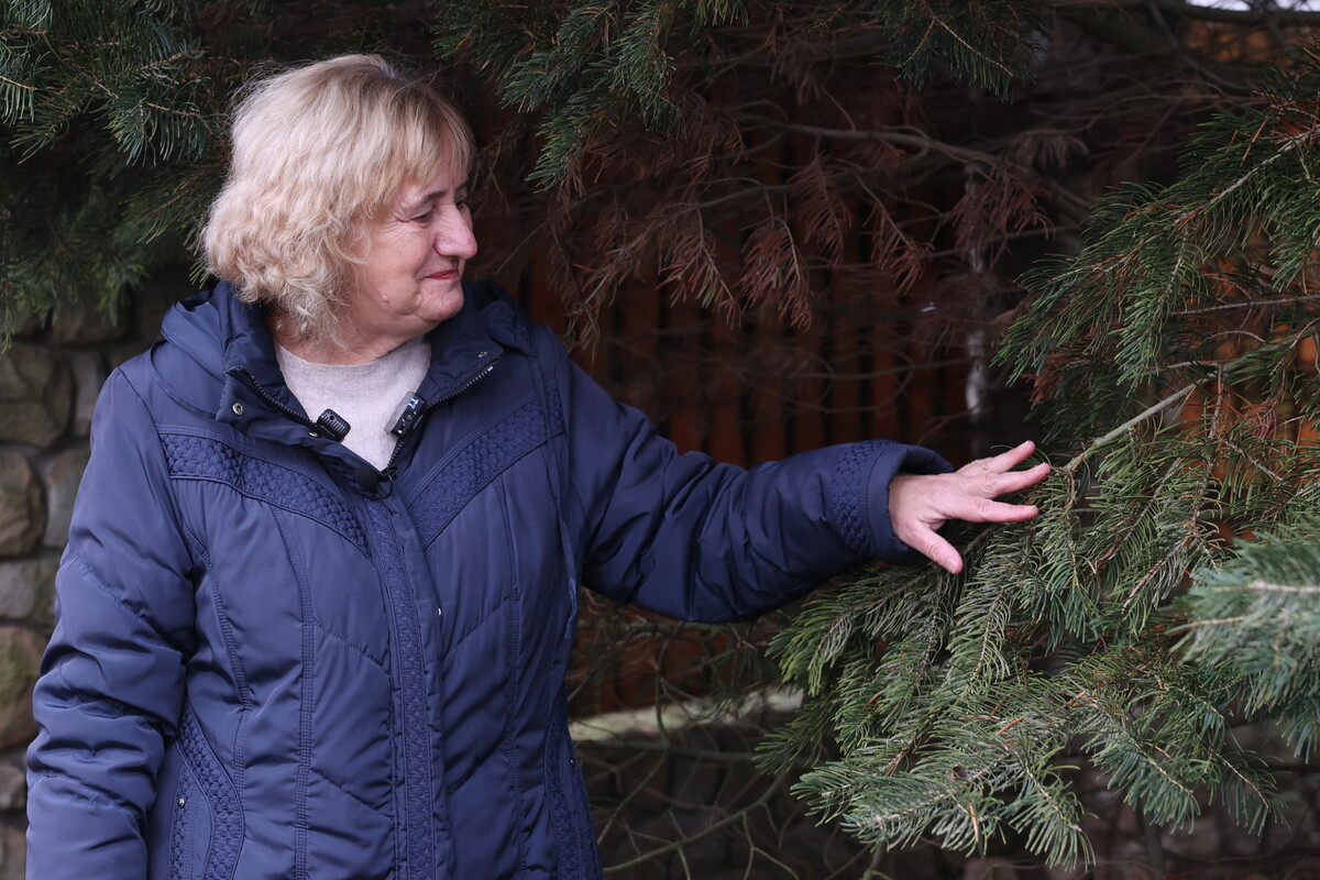 Любов Пашечко, простягнувши праву руку до гілок хвойного дерева, зі сумом і ніжністю дивиться на них.
